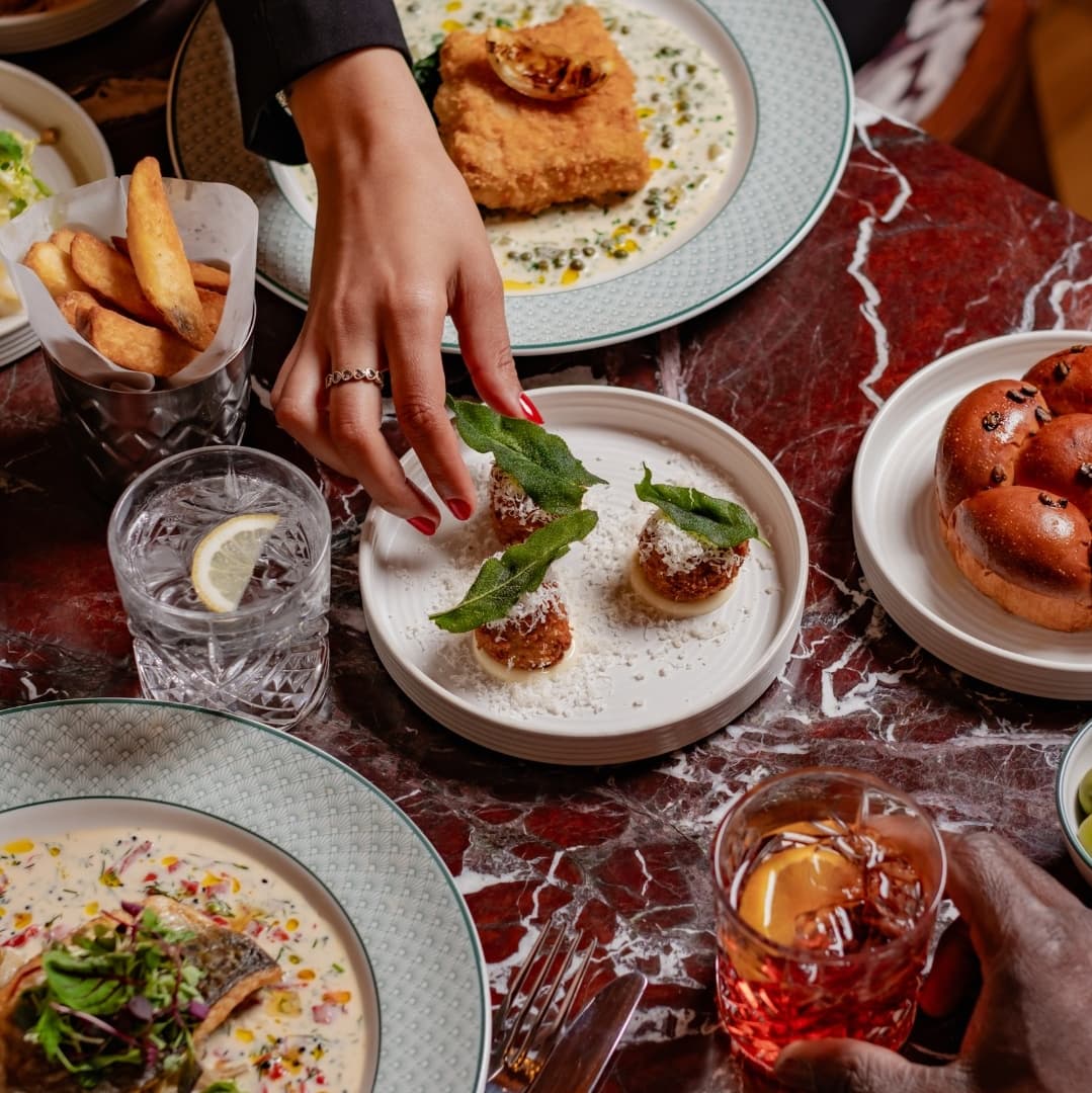 Artfully Plated Dishes with Fresh Herbs A hand reaches for a plate of garnished appetizers, surrounded by vibrant dishes and drinks on a marble table.