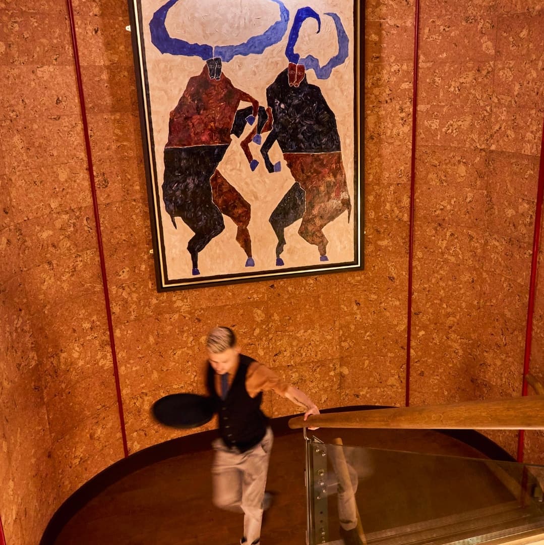 Dynamic Artwork Above Spiraling Staircase A server descends a wooden staircase, carrying a tray, with a large abstract bull painting on the wall above.