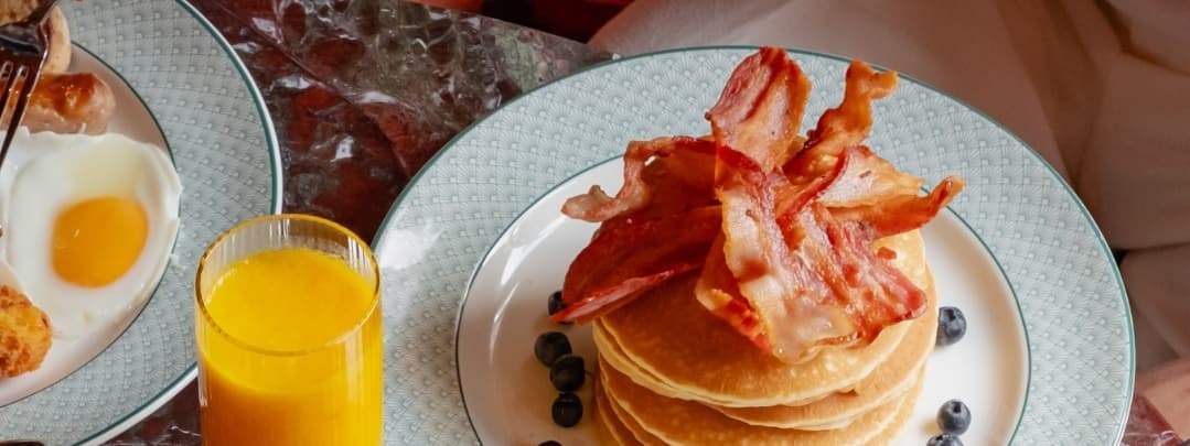 Stack of fluffy pancakes topped with crispy bacon, blueberries, and a glass of orange juice on a stylish table.
