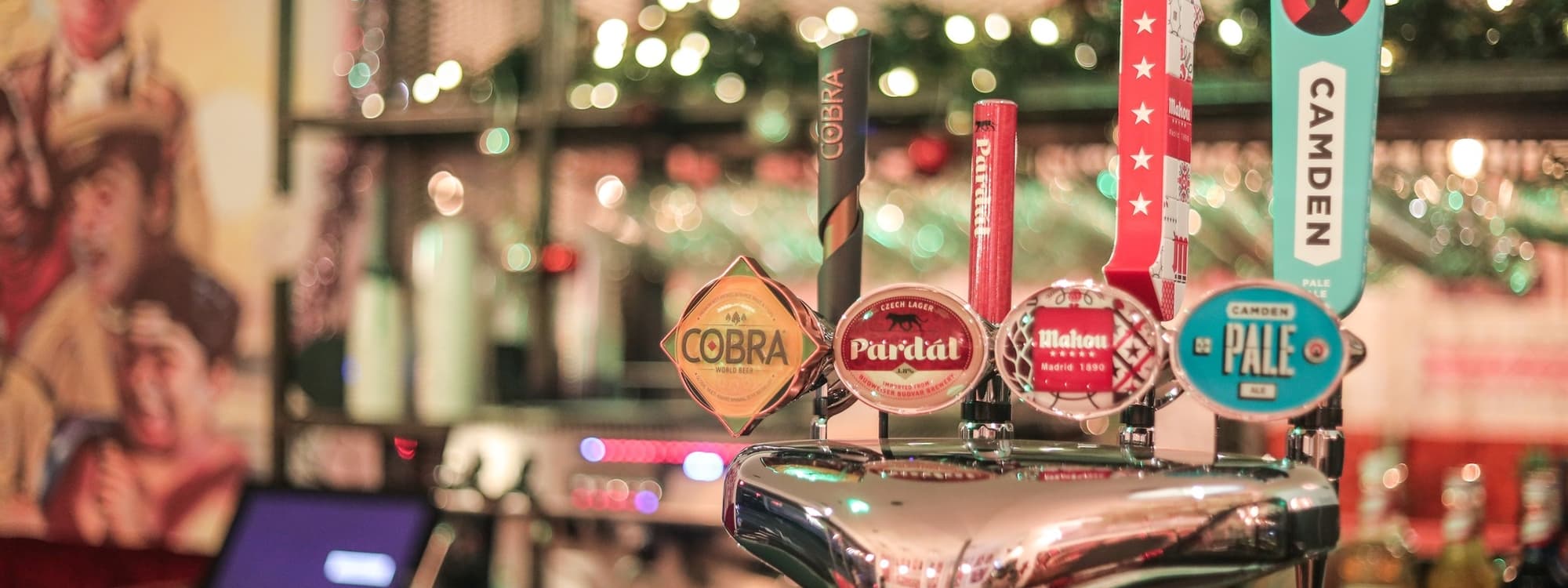 Colorful beer taps with various labels, set against a festive backdrop of twinkling lights and vibrant decor.