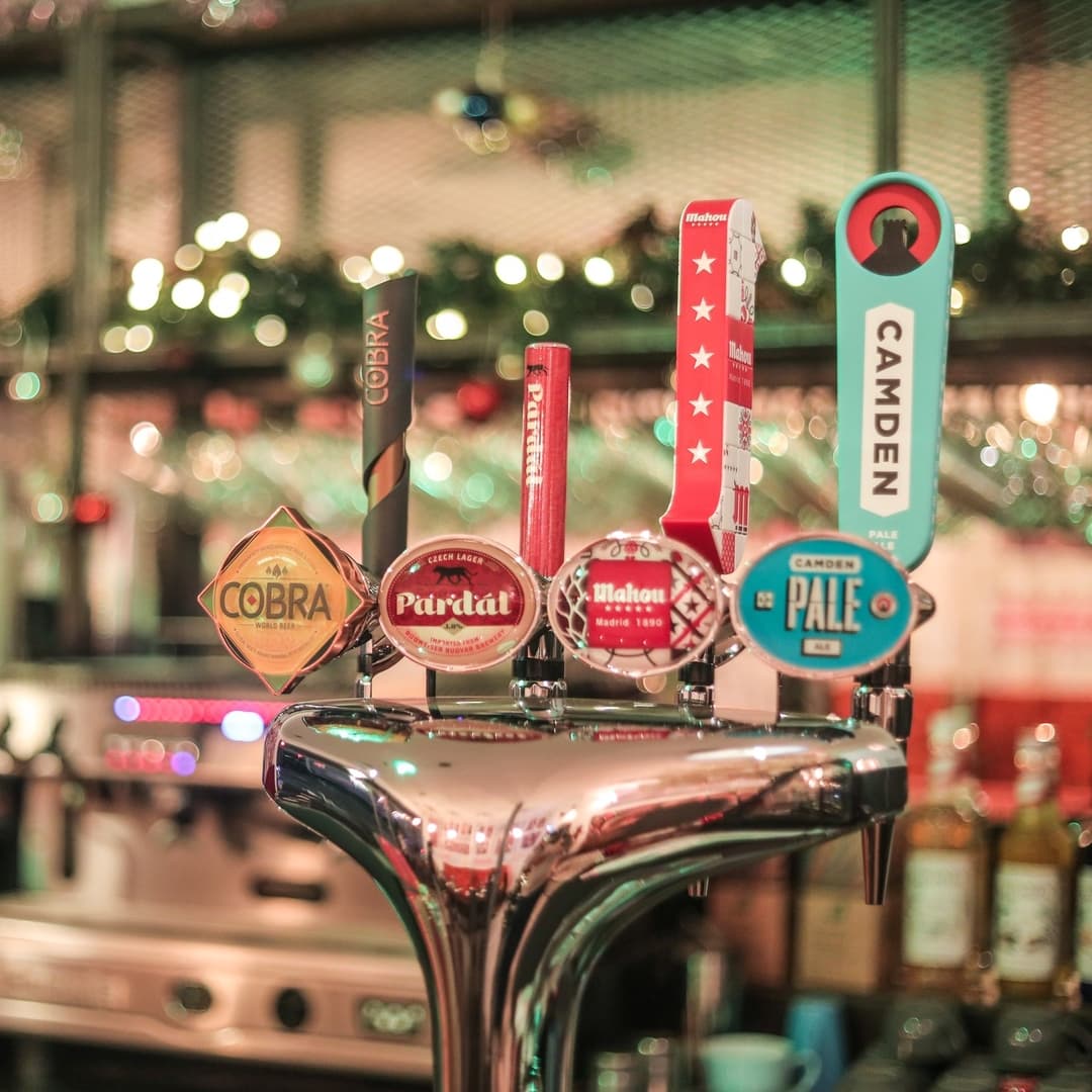 Colorful beer taps lined up on a polished chrome bar, with festive lights creating a warm, inviting atmosphere.