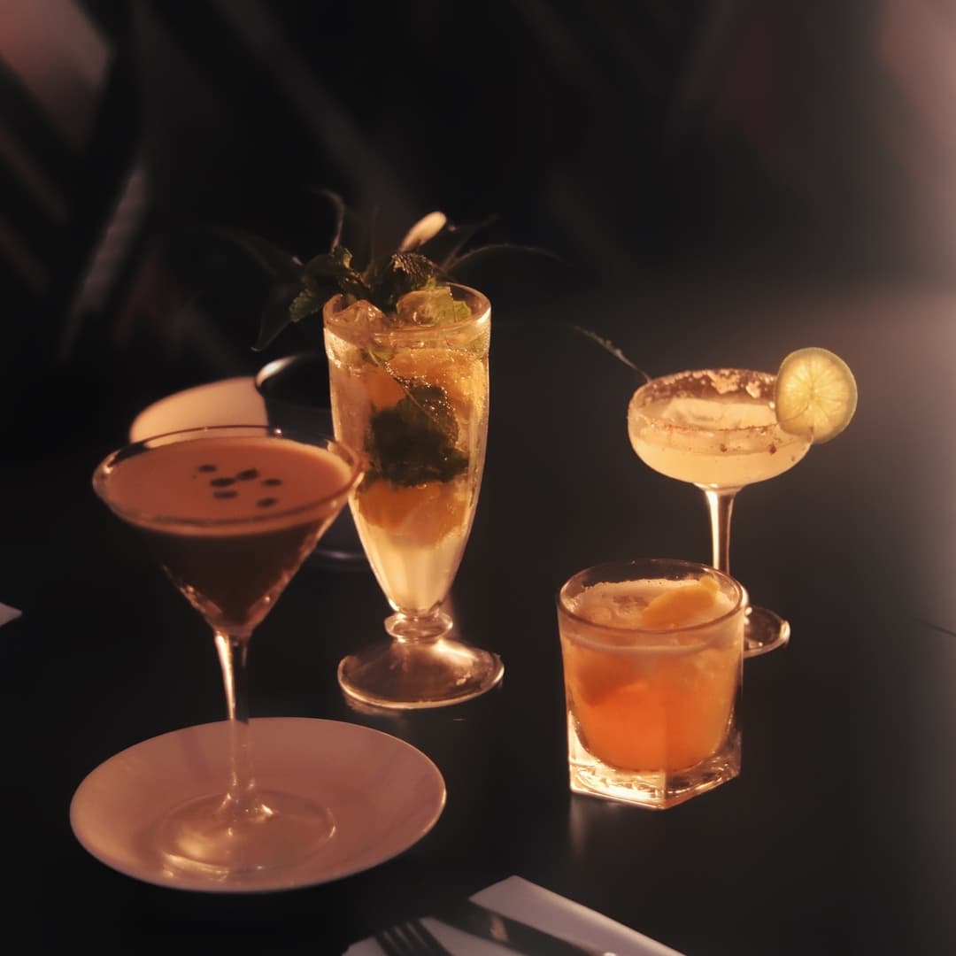 Four colorful cocktails on a dark table, featuring garnishes of fruit and herbs, creating a vibrant, inviting atmosphere.