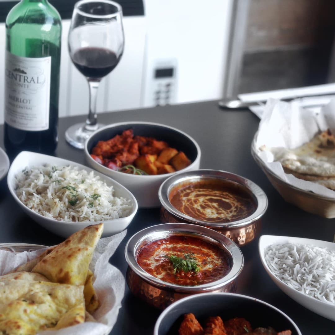 A table set with vibrant Indian dishes, rice, naan, and a glass of red wine, creating a warm dining atmosphere.