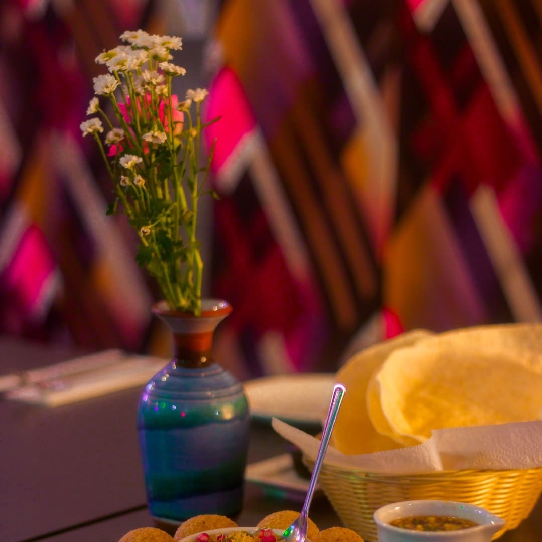 Colorful table setting with a small vase of flowers, traditional Indian dishes, and vibrant patterned backdrop.