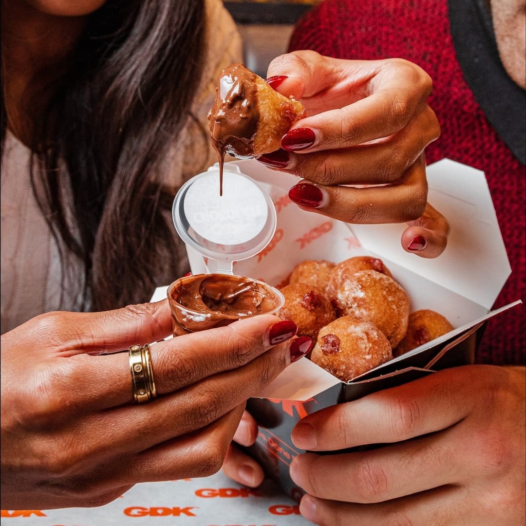 Two hands reach for a chocolate-dipped donut ball from a GDK takeout box, showcasing a cozy dining atmosphere.