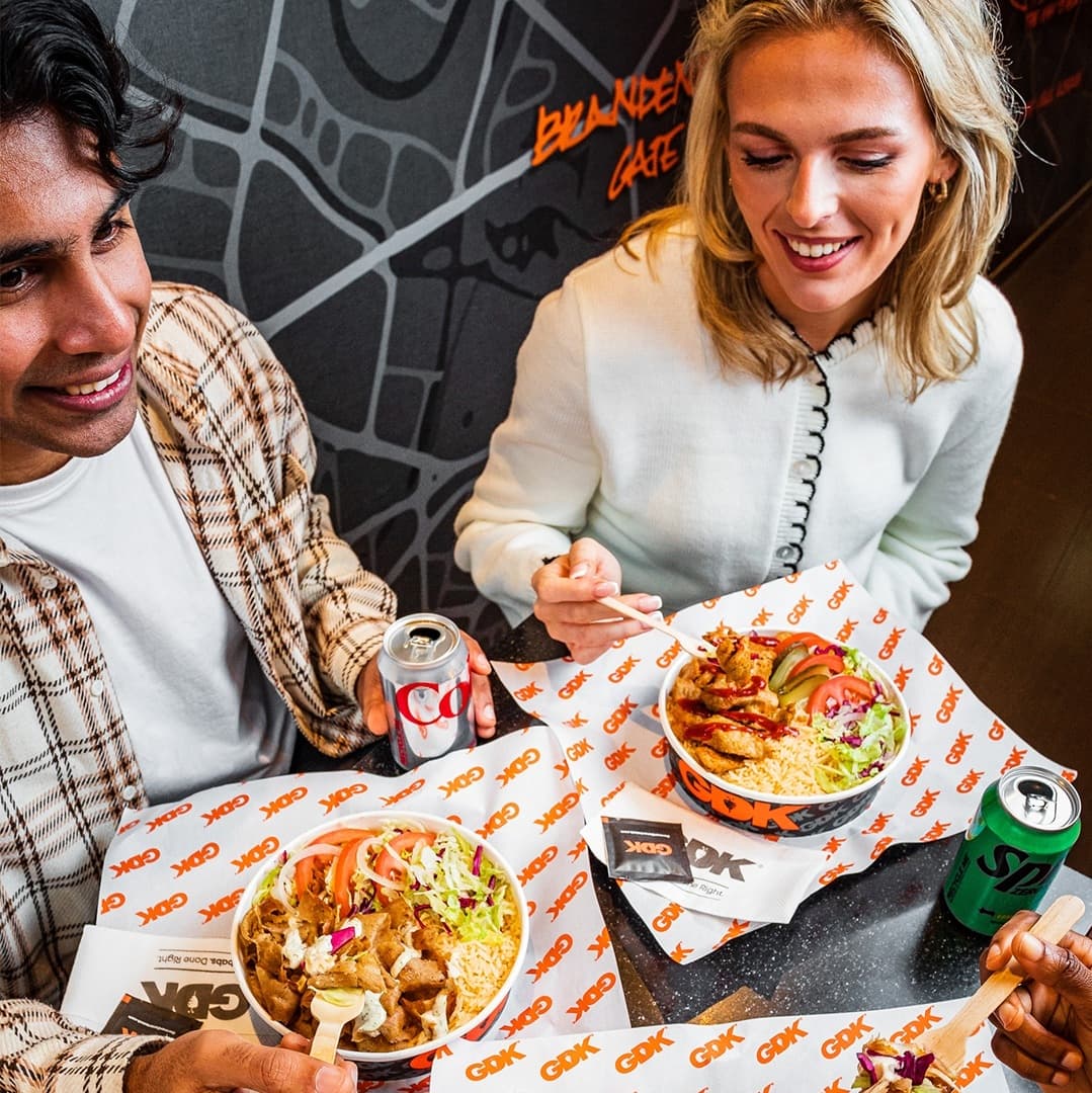 Two diners enjoy colorful bowls of kebab with fresh vegetables, accompanied by soft drinks, in a vibrant restaurant setting.
