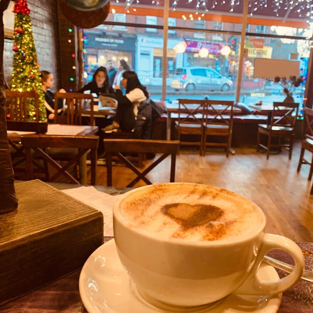 A cozy café scene featuring a heart-shaped cappuccino, wooden tables, and festive decorations with twinkling lights.