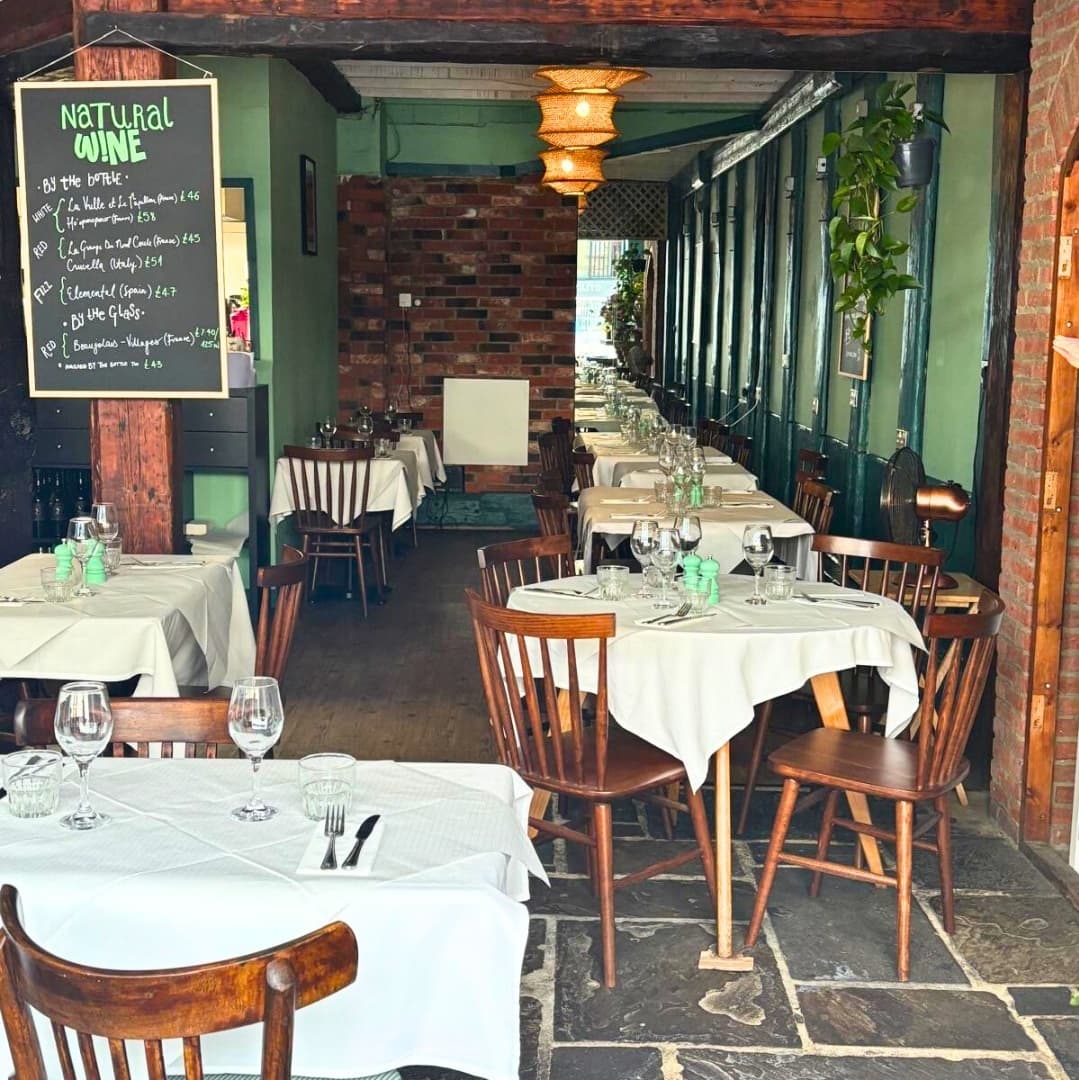Brightly lit bistro with wooden beams, white tablecloths, and green accents, creating a cozy, inviting dining atmosphere.