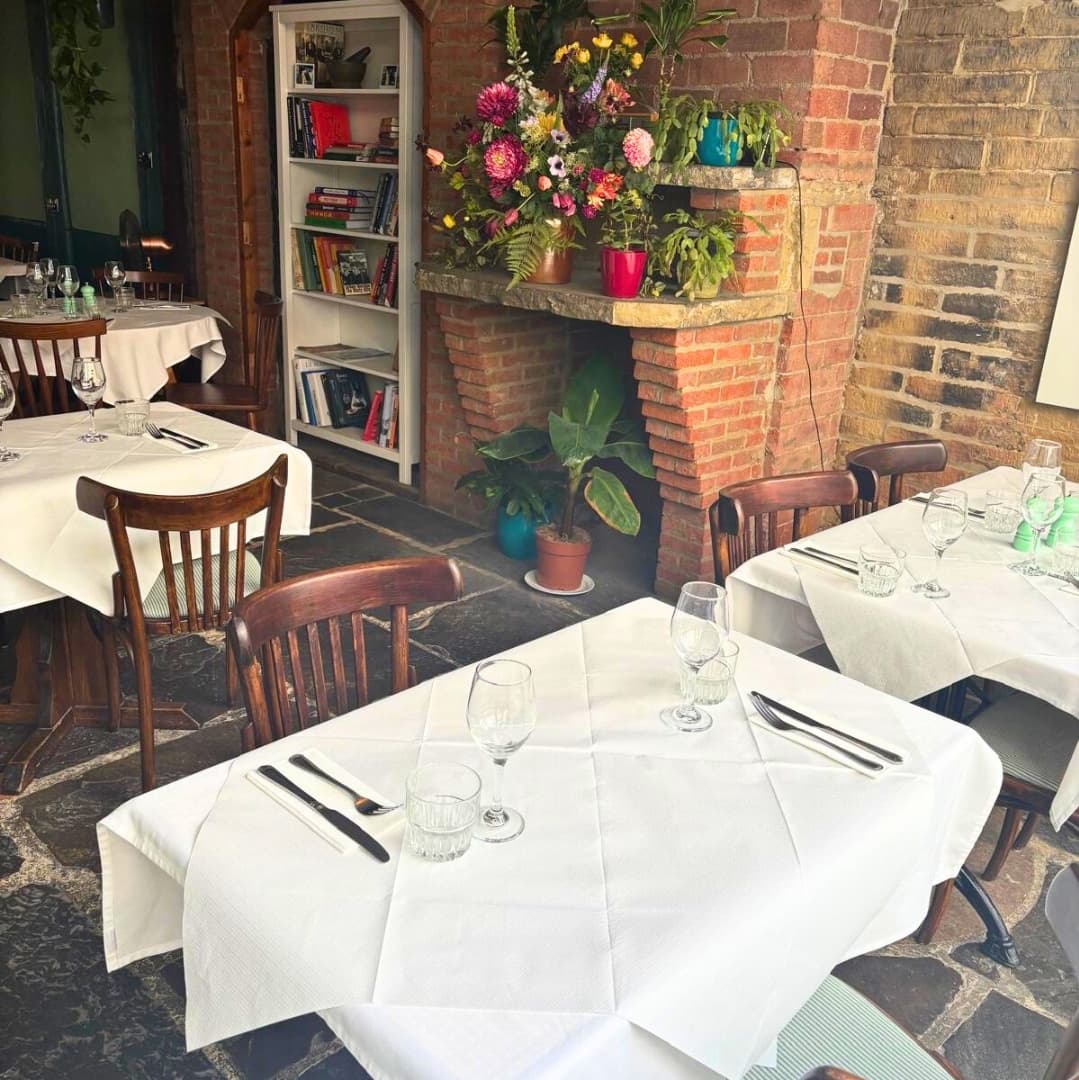 Cozy bistro interior with wooden tables, white tablecloths, and vibrant floral arrangements against a brick wall.