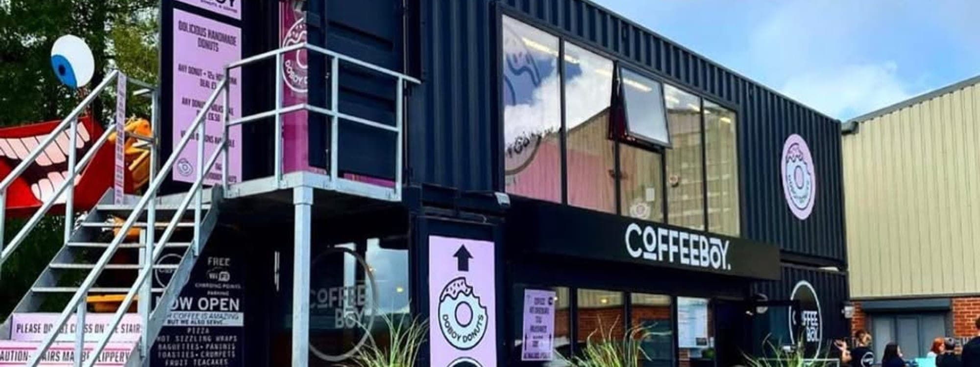 Coffeeboy Headingley exterior during the day, showing the black shipping container building with the Coffeeboy logo and outdoor seating area