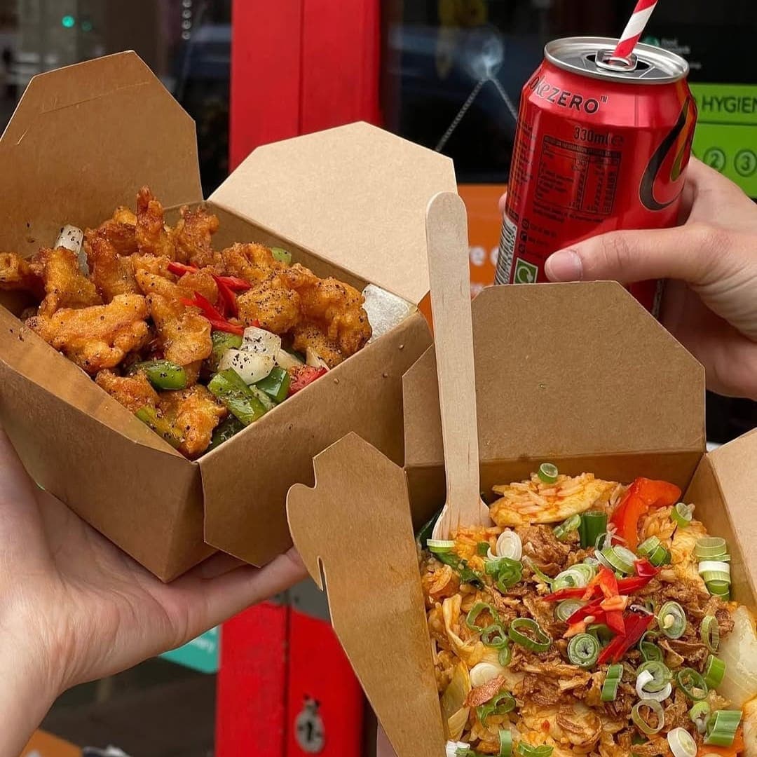 Two Sri Non Thai Street Food takeaway boxes with fried chicken and rice held up outside the restaurant