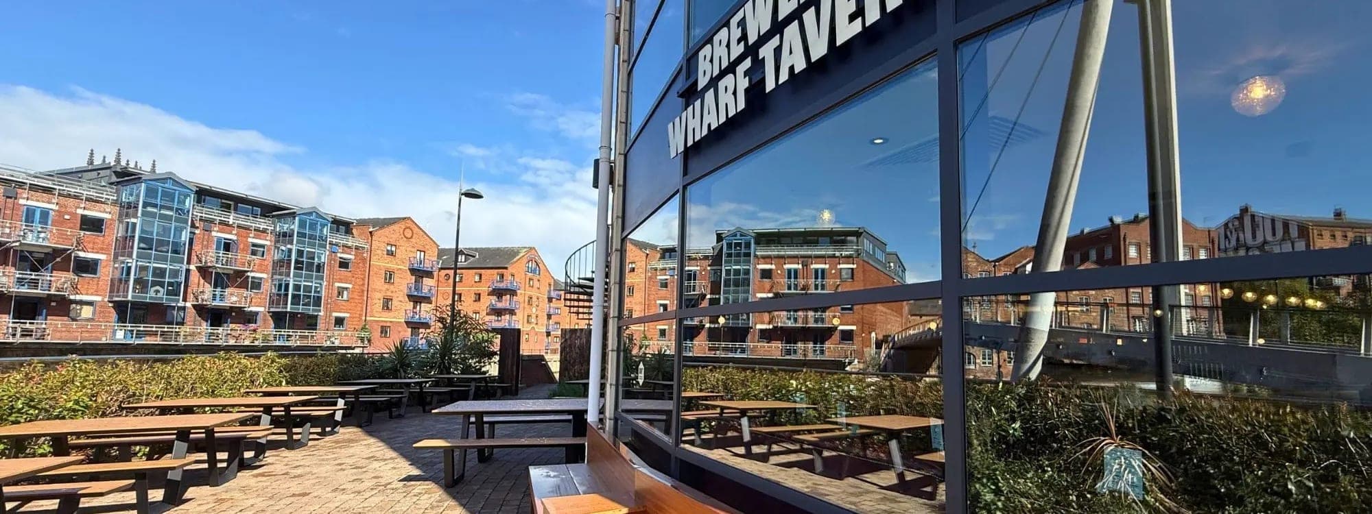 Outdoor beer terrace at Brewery Wharf Tavern with picnic benches beside the waterfront and the venue signage visible on the glass facade