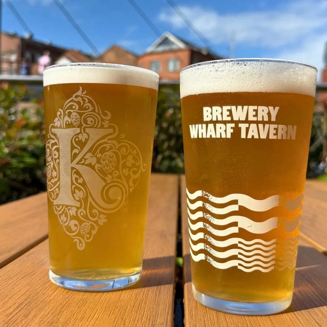 Two pints of golden lager on a wooden table on the outdoor terrace at Brewery Wharf Tavern with blue sky behind