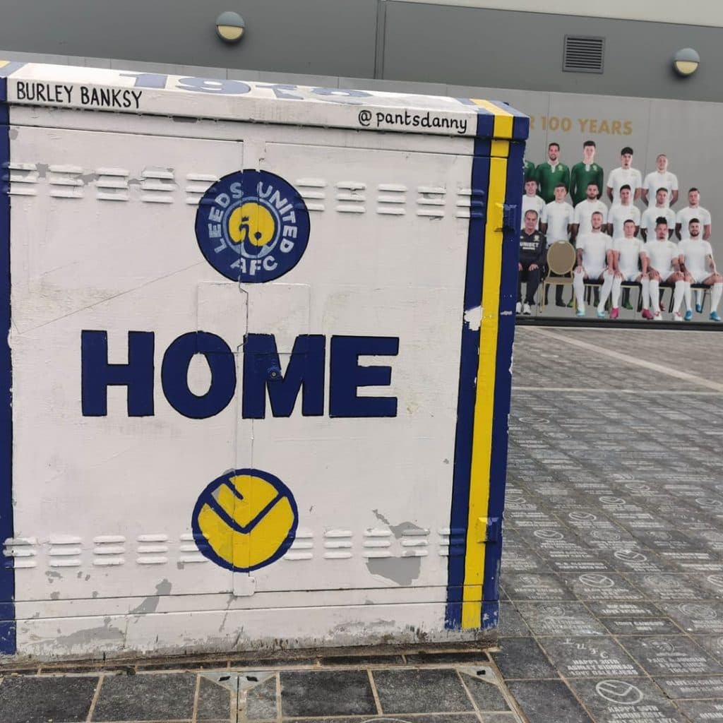 Burley Banksy painted box at Centenary Square, Elland Road — "HOME" and the LUFC badge in club colours