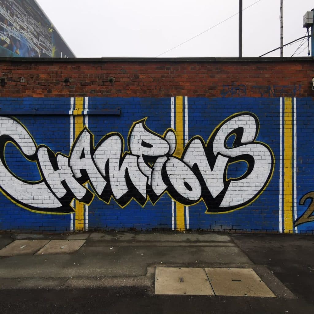 Champions mural by Mateusz Klich and Adam Duffield (Meds One) on the Lowfields Road wall, Elland Road, Leeds — graffiti-style "Champions 2020" lettering with trophy