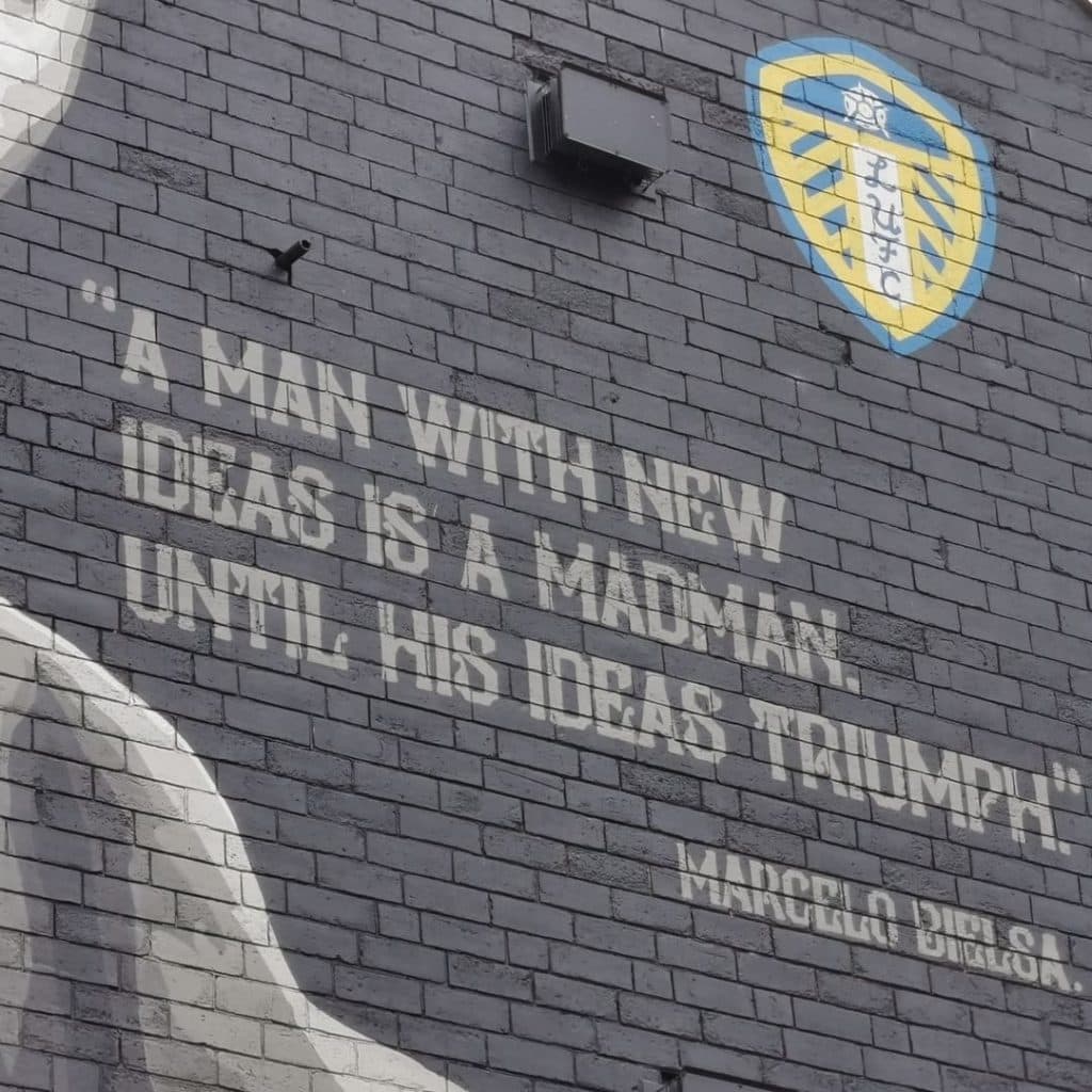 Marcelo Bielsa mural by Tankpetrol on the side gable of One Stop Stores, Hyde Park Road, Leeds — black-and-white portrait with the quote "A man with new ideas is a madman until his ideas triumph"