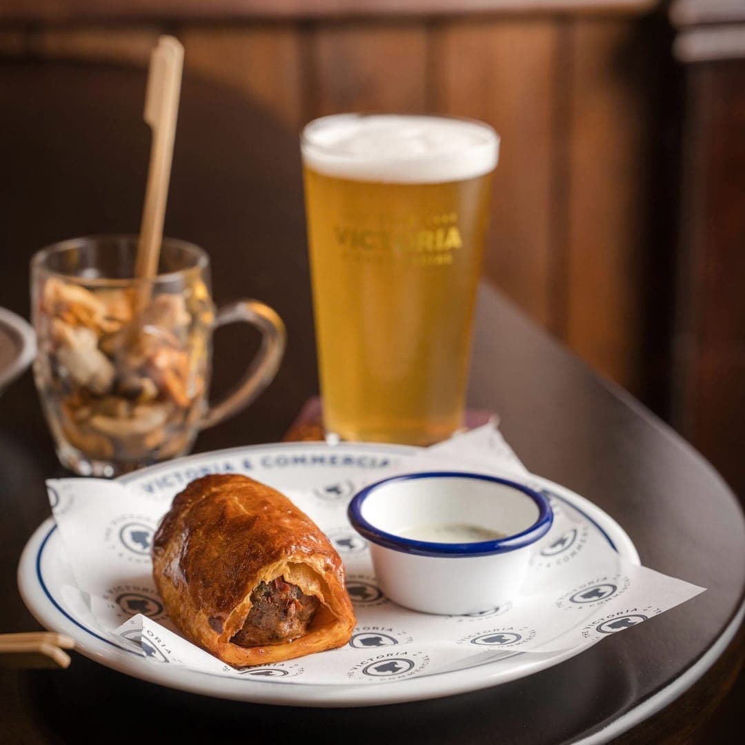 A branded Victoria pint of golden lager with a sausage roll on a branded Victoria and Commercial plate and mixed nuts in a glass mug