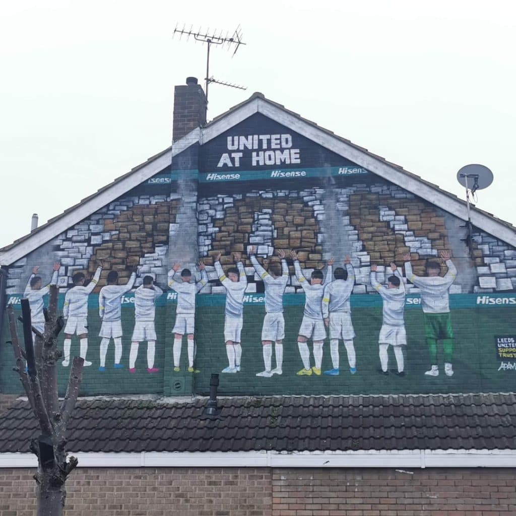 United at Home mural by Adam Duffield on a private house gable at Wesley Street and Hoxton Mount, Leeds — team line-up with arms raised in front of an Elland Road backdrop