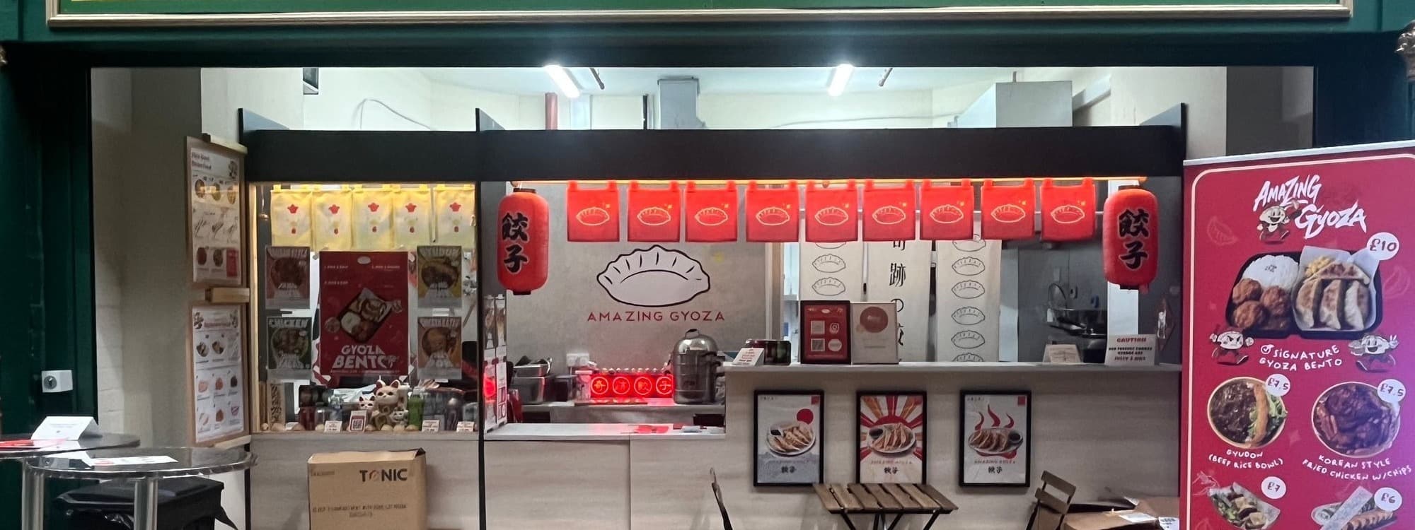 The Amazing Gyoza stall inside Leeds Kirkgate Market, with red paper lanterns, a gyoza wall logo, menu posters and a branded standee showing signature dishes.