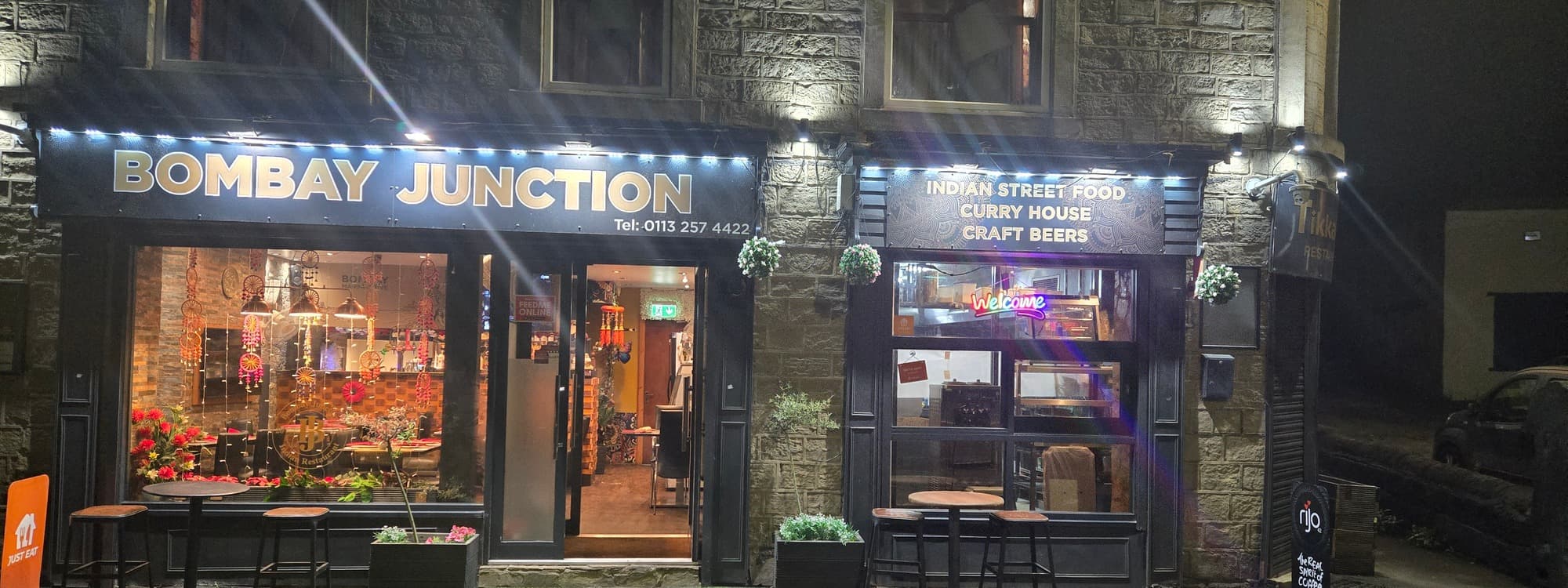 The Bombay Junction restaurant in Bramley, Leeds, at night, with a BOMBAY JUNCTION sign on the left, INDIAN STREET FOOD / CURRY HOUSE / CRAFT BEERS sign on the right, Yorkshire stone facade, and outside seating with high tables and stools.