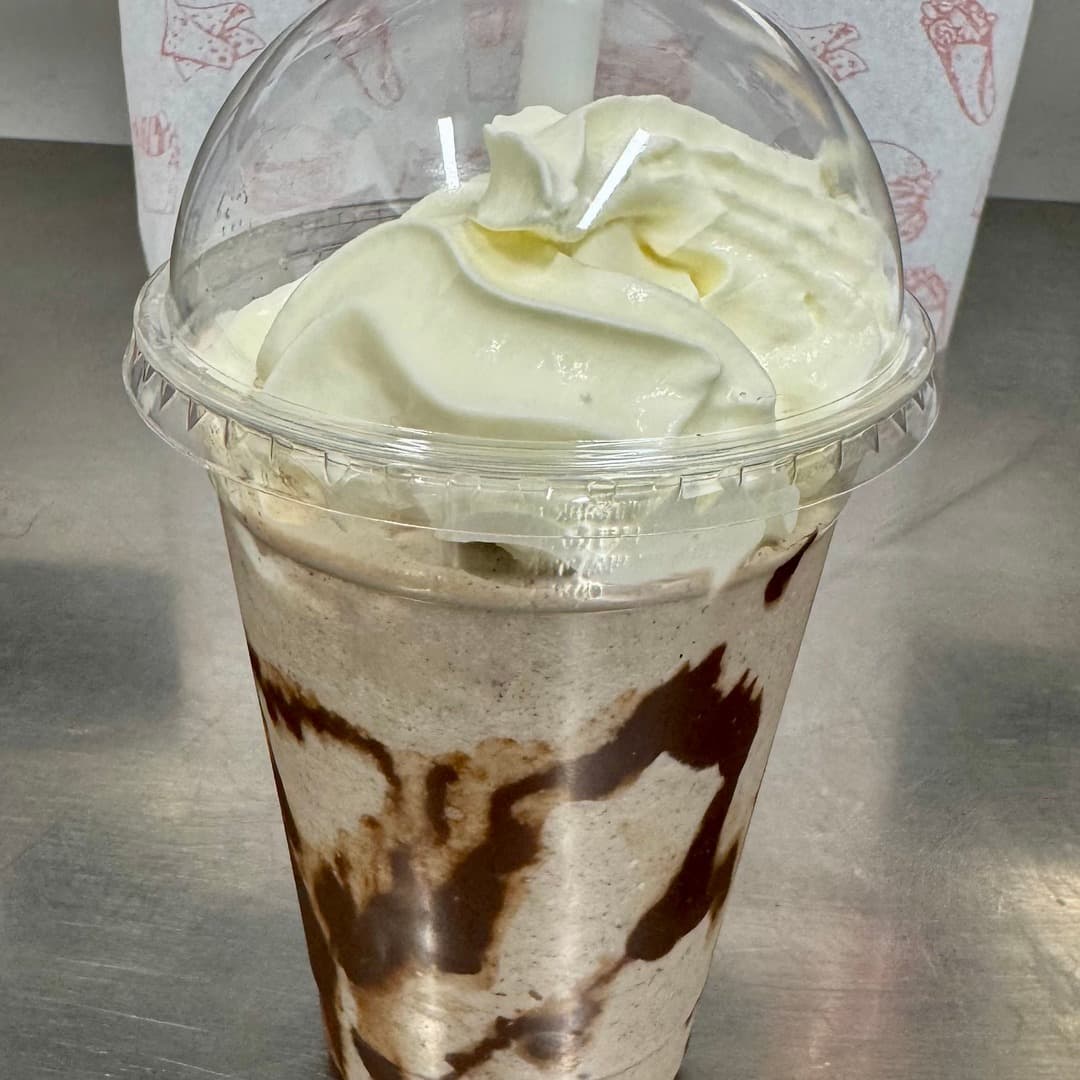 A milkshake with whipped cream piled high and chocolate sauce drizzled inside the cup, on a stainless steel counter at Fatbros, Bramley, Leeds.