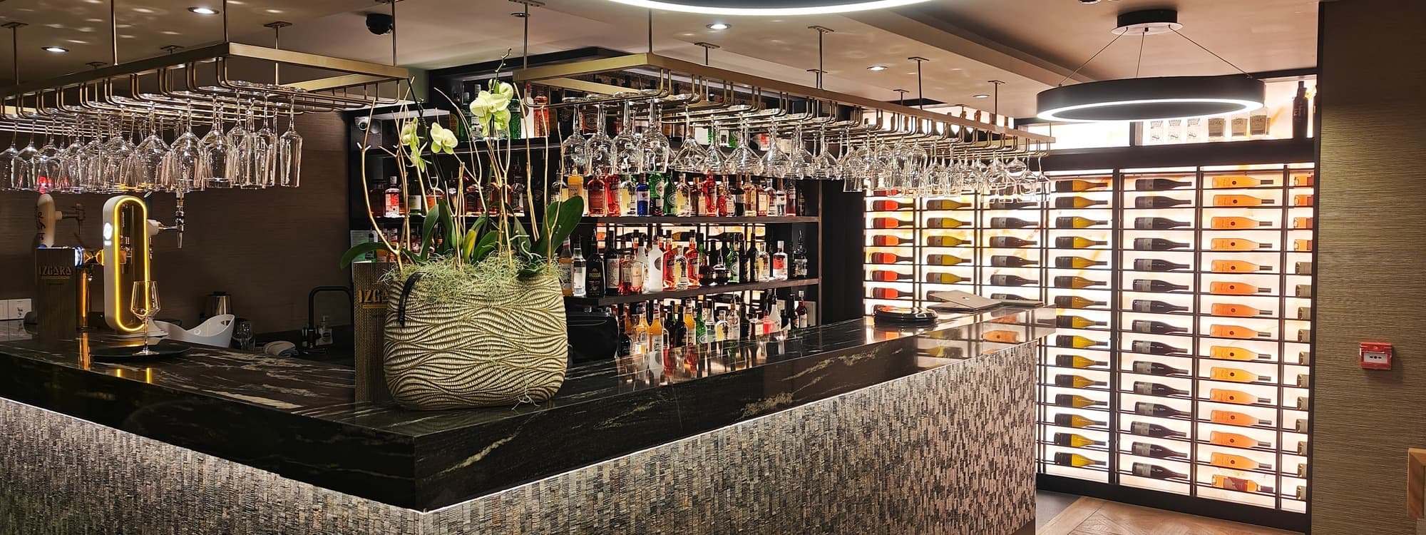 The bar at Izgara Mediterranean Grill in Garforth, Leeds, with a marble counter, mosaic-tiled bar front, hanging glassware, spirits shelves, illuminated wine wall and circular LED ring lights overhead.
