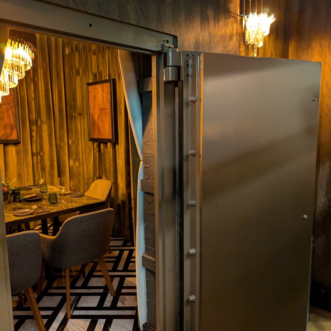The original bank vault door at Izgara Mediterranean Grill in Garforth, Leeds, opening onto a private dining room with gold curtains, monochrome patterned floor and a chandelier — a heritage feature from the building's former life as a bank.