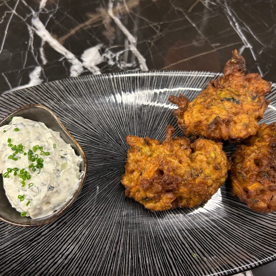 Three golden courgette fritters served with a small bowl of herbed yogurt topped with chives, on a striped black plate at Izgara Mediterranean Grill, Garforth, Leeds.