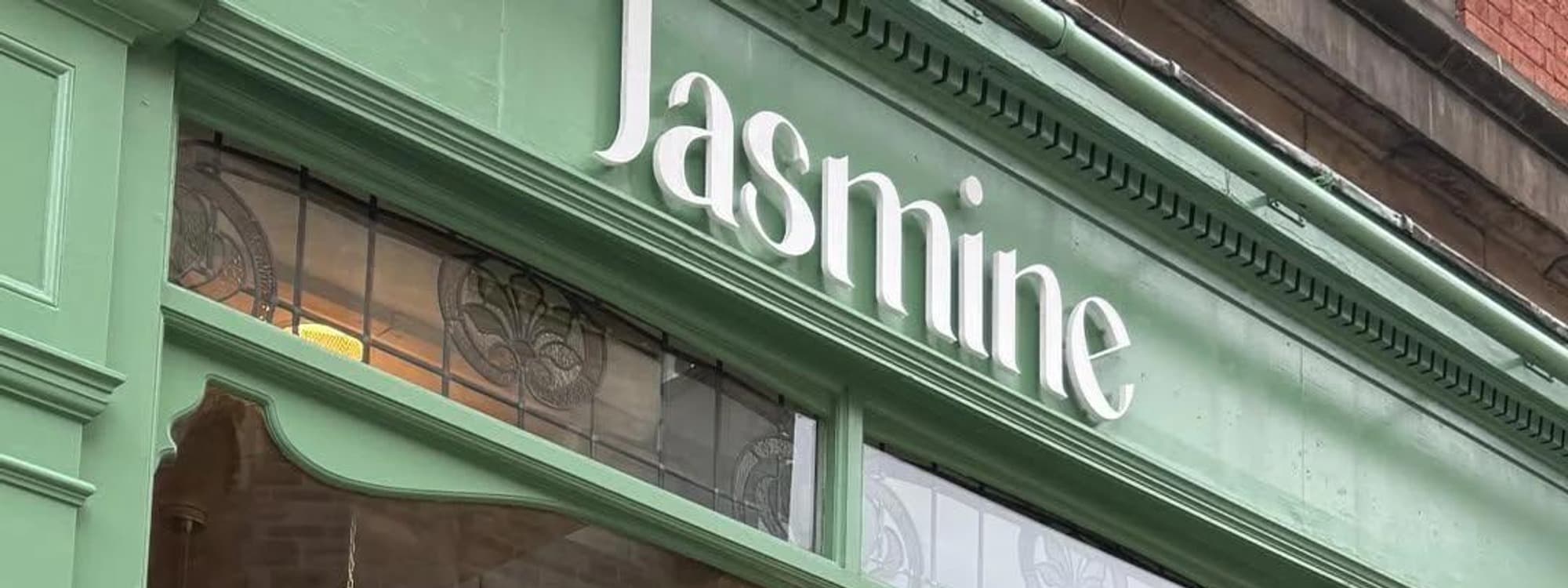 The pale-green painted shopfront of Jasmine Cafe in Leeds, with the white Jasmine wordmark on the fascia and decorative leaded glass panels below.