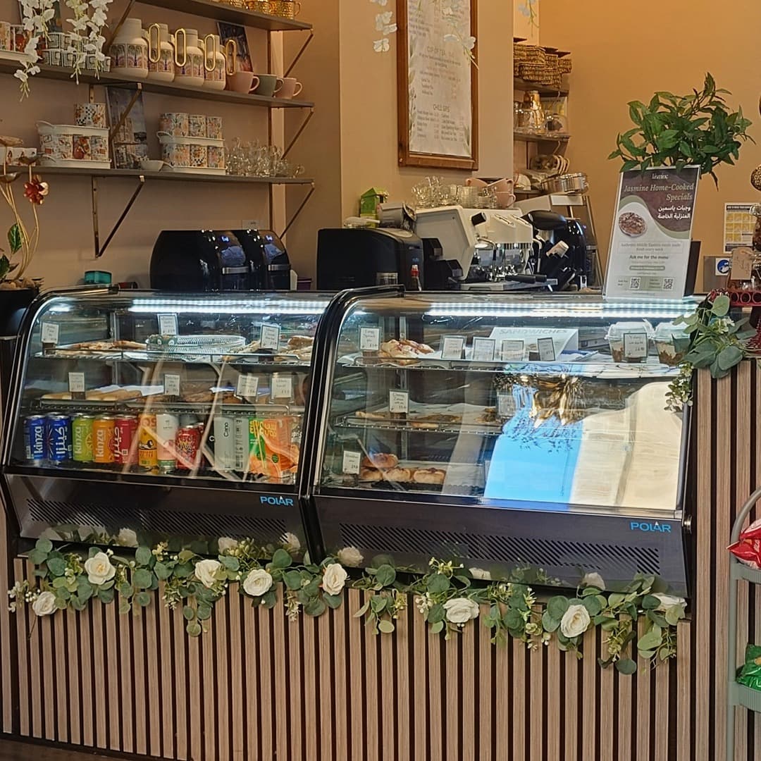 The counter at Jasmine Cafe, Leeds, with refrigerated display cases of pastries and drinks, and decorative Arabic mugs and teapots on shelves above.