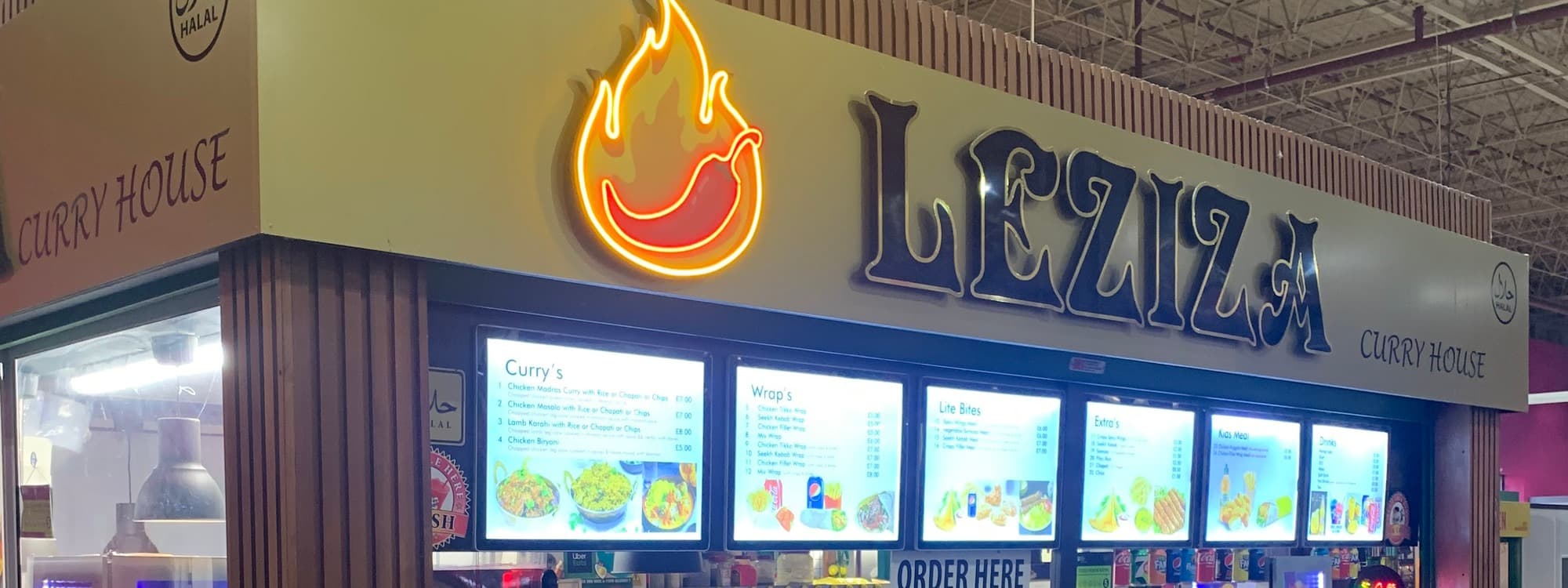 The Leziza Curry House stall in Leeds Kirkgate Market, with a neon-lit chili-flame logo, gold LEZIZA wordmark and CURRY HOUSE subtitle, illuminated menu boards for curries, wraps, and lite bites, and an ORDER HERE counter sign.