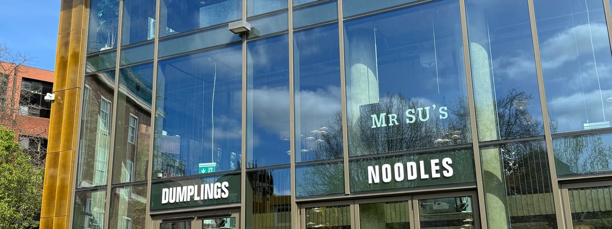 The glass facade of Mr Su Noodles on Cross Belgrave Street, Leeds, with MR SU'S DUMPLINGS NOODLES signage and the modern brass-trimmed Vita Student building behind.
