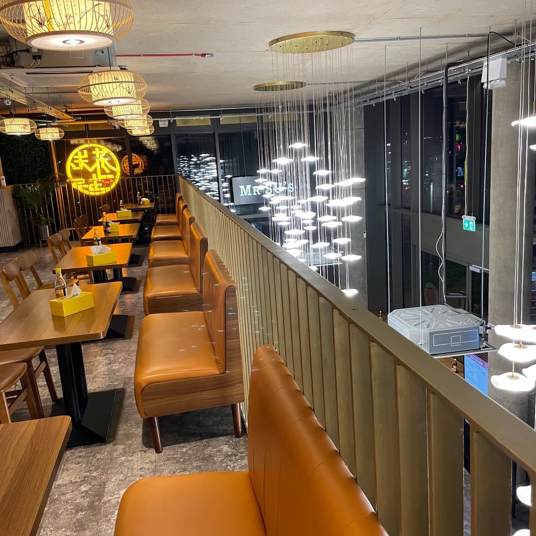 The dining room at Mr Su Noodles, Leeds, showing tan leather banquettes, wooden tables, bamboo pendant lamps, neon Chinese signage, and a cascading butterfly-light installation.