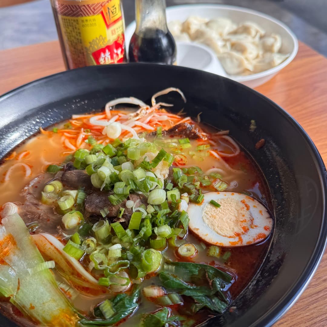 A bowl of spicy beef noodles with marinated egg, pak choi, spring onions, and a side of dumplings at Mr Su Noodles, Leeds.
