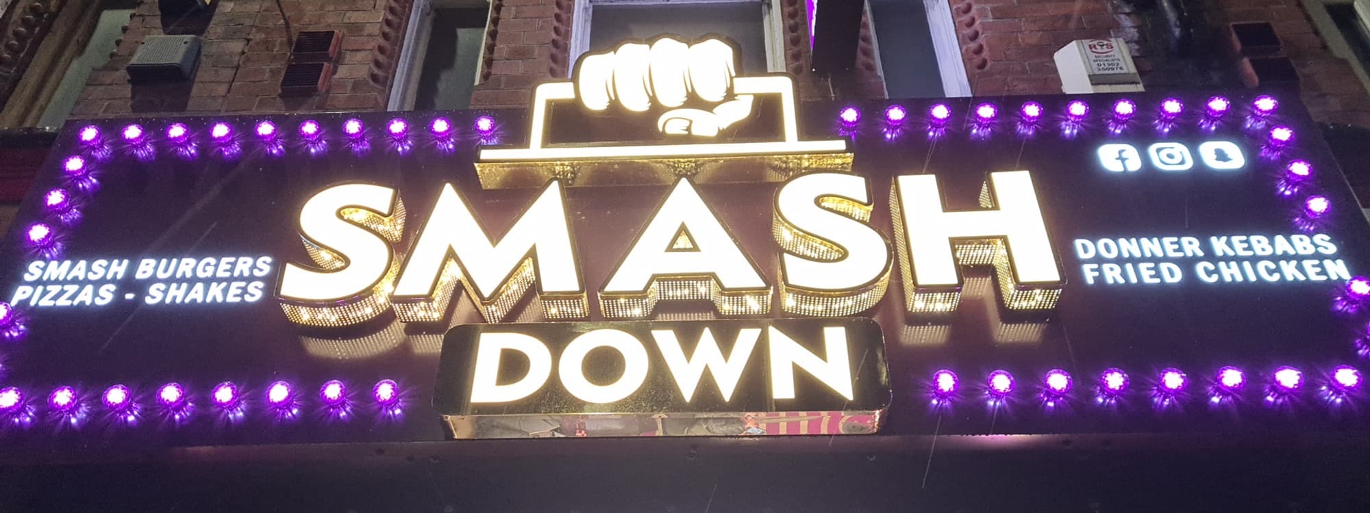 The neon SMASH DOWN sign on Woodhouse Lane in Leeds, with a fist logo above the brand name and purple bulb borders. Side text reads SMASH BURGERS PIZZAS SHAKES on the left and DONNER KEBABS FRIED CHICKEN on the right.