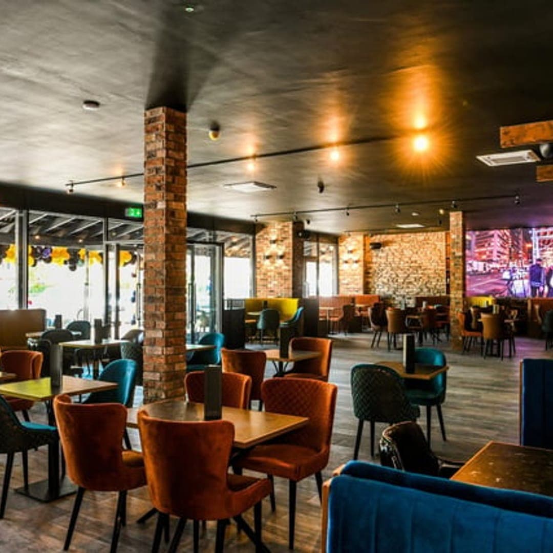 Spacious dining room interior at TAPAS BAR by olive branch with exposed brick pillars, coloured velvet chairs and large screens at the back