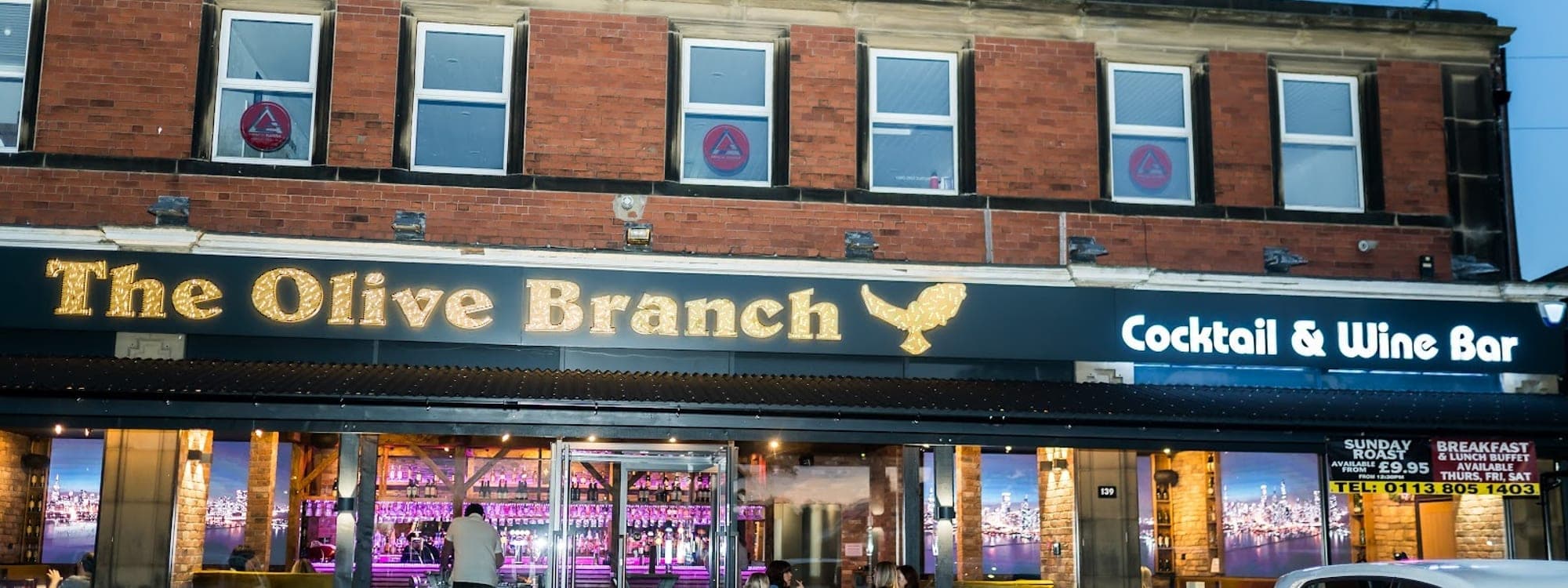 The Olive Branch building at night with illuminated gold signage and Cocktail & Wine Bar sign, guests seated outside on Street Lane Roundhay Leeds