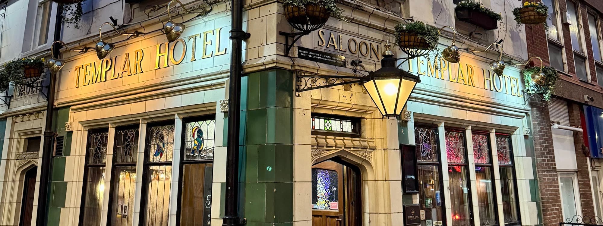 The dusk exterior of the Templar pub on Templar Street, Leeds, with green and white glazed tiles, gold TEMPLAR HOTEL lettering on both sides of the corner, a SALOON arch over the entrance, and a glowing brass street lamp.