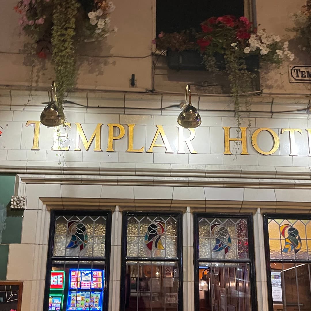 Close-up of the gold TEMPLAR HOTEL lettering on white glazed tiles, with hanging flower baskets above and stained-glass shopfront windows below, on the Templar pub in Leeds.
