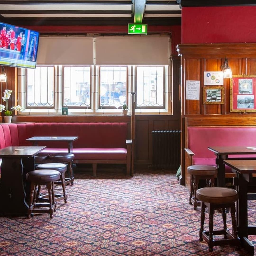 The interior of the Templar pub, Leeds, with red velvet banquettes, dark wood panelling, leaded stained-glass windows, patterned carpet, and a TV showing live sport.