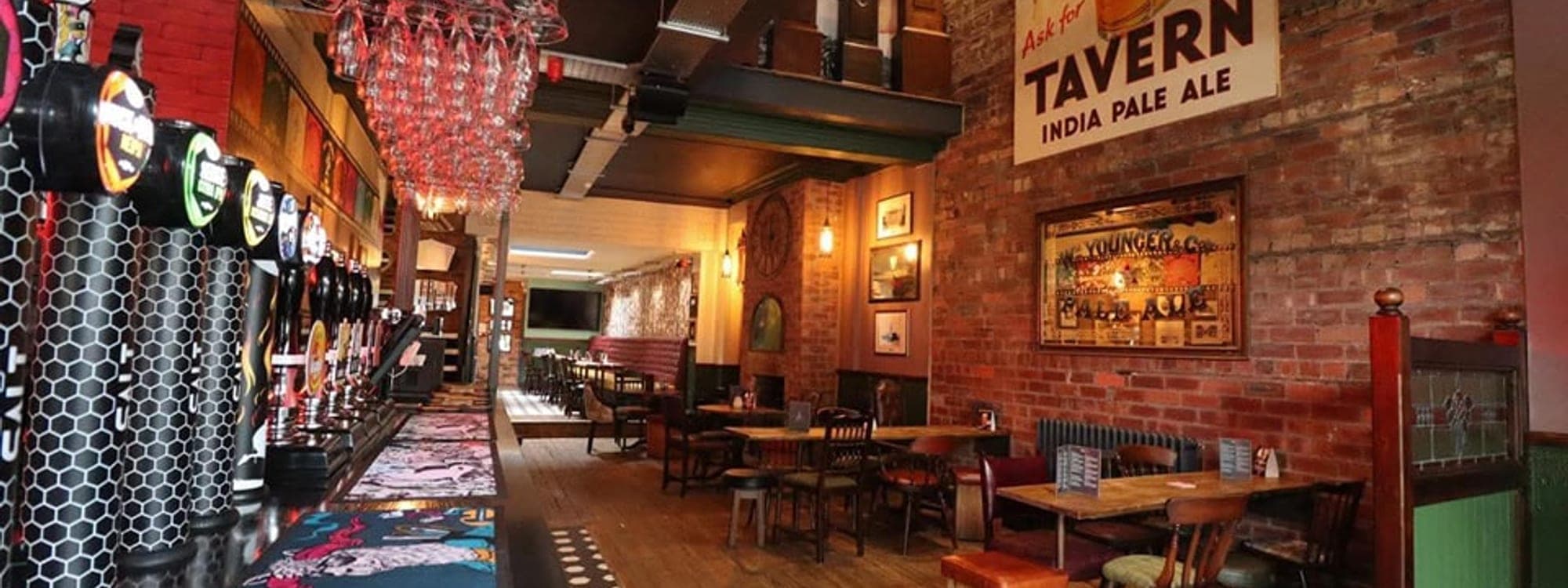 Wide interior shot of The Clock Inn showing the bar with multiple ale taps, glass chandelier, brick walls and vintage TAVERN India Pale Ale sign