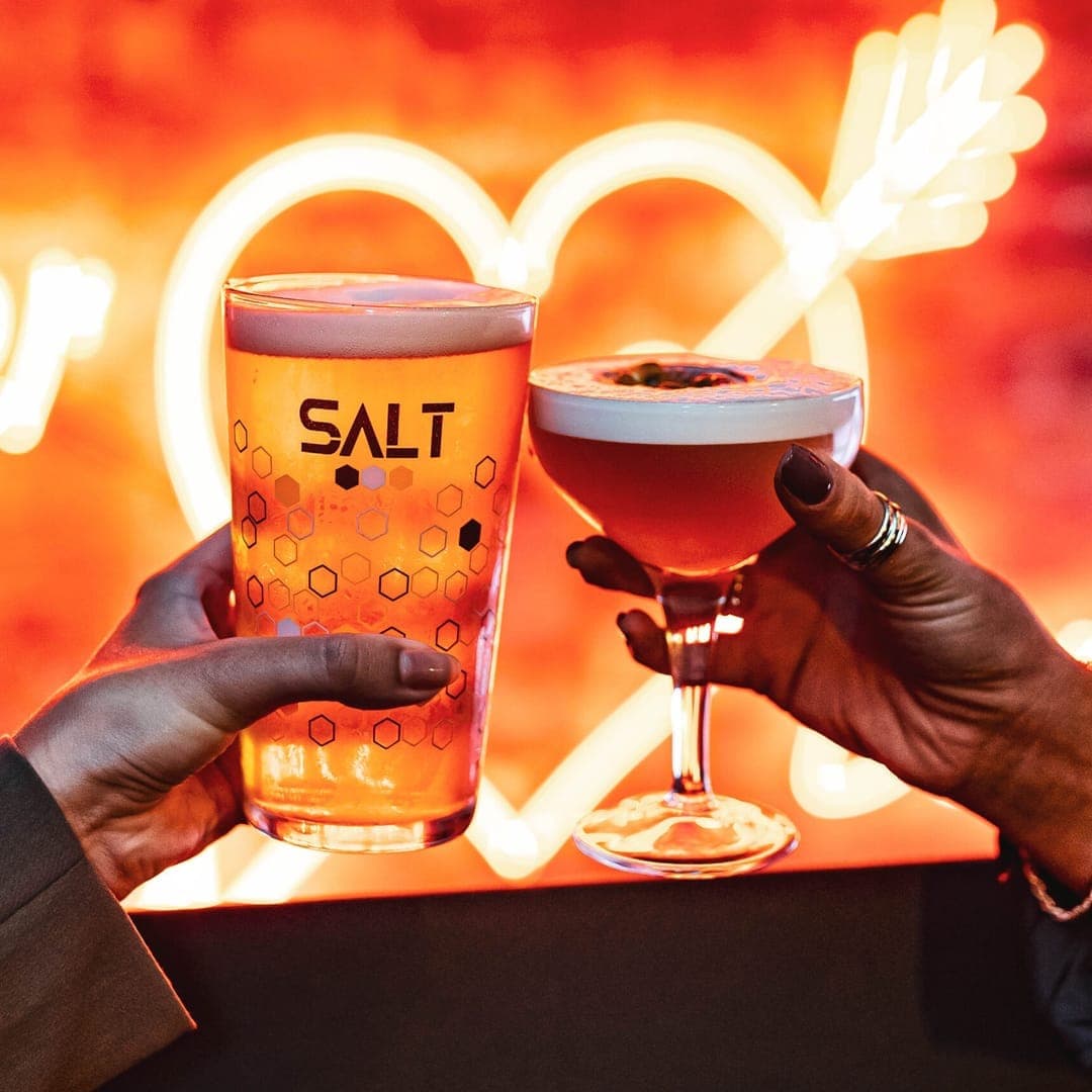 Two hands raising a SALT-branded pint of lager and a coupe cocktail in front of a glowing orange neon heart sign