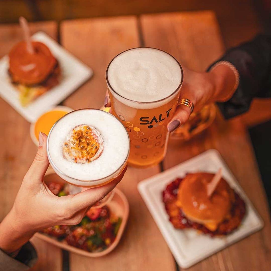 Overhead shot of two hands raising a passionfruit cocktail and a SALT pint of beer over a table with burgers and sides