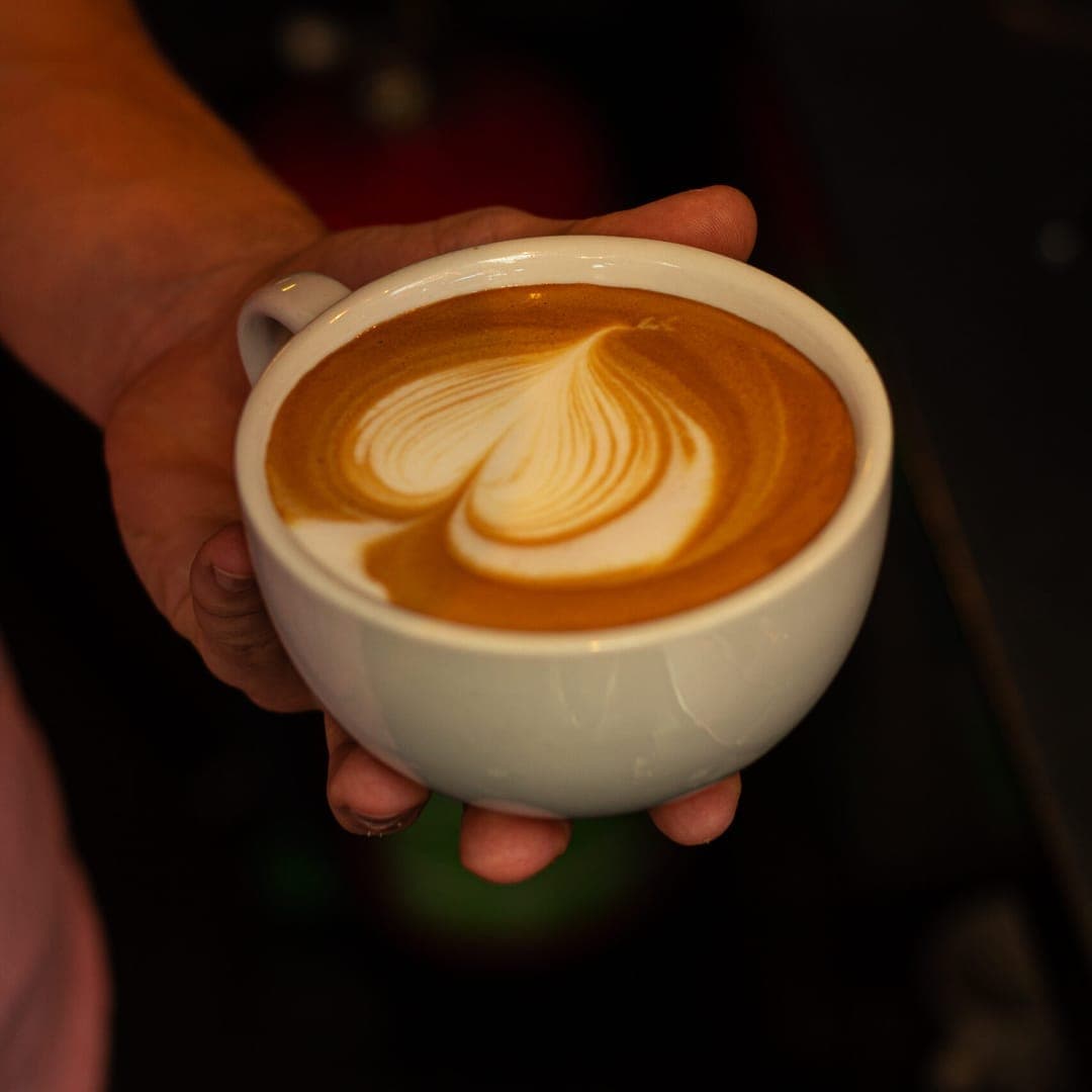 Hand holding a white ceramic cup of flat white coffee with intricate heart latte art on a dark background