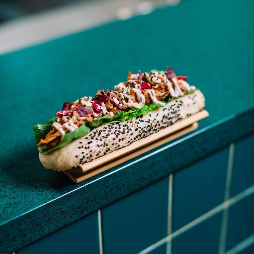 Sesame-seeded hot dog bun filled with pulled meat, lettuce, sauce and microgreens resting on a teal tiled surface