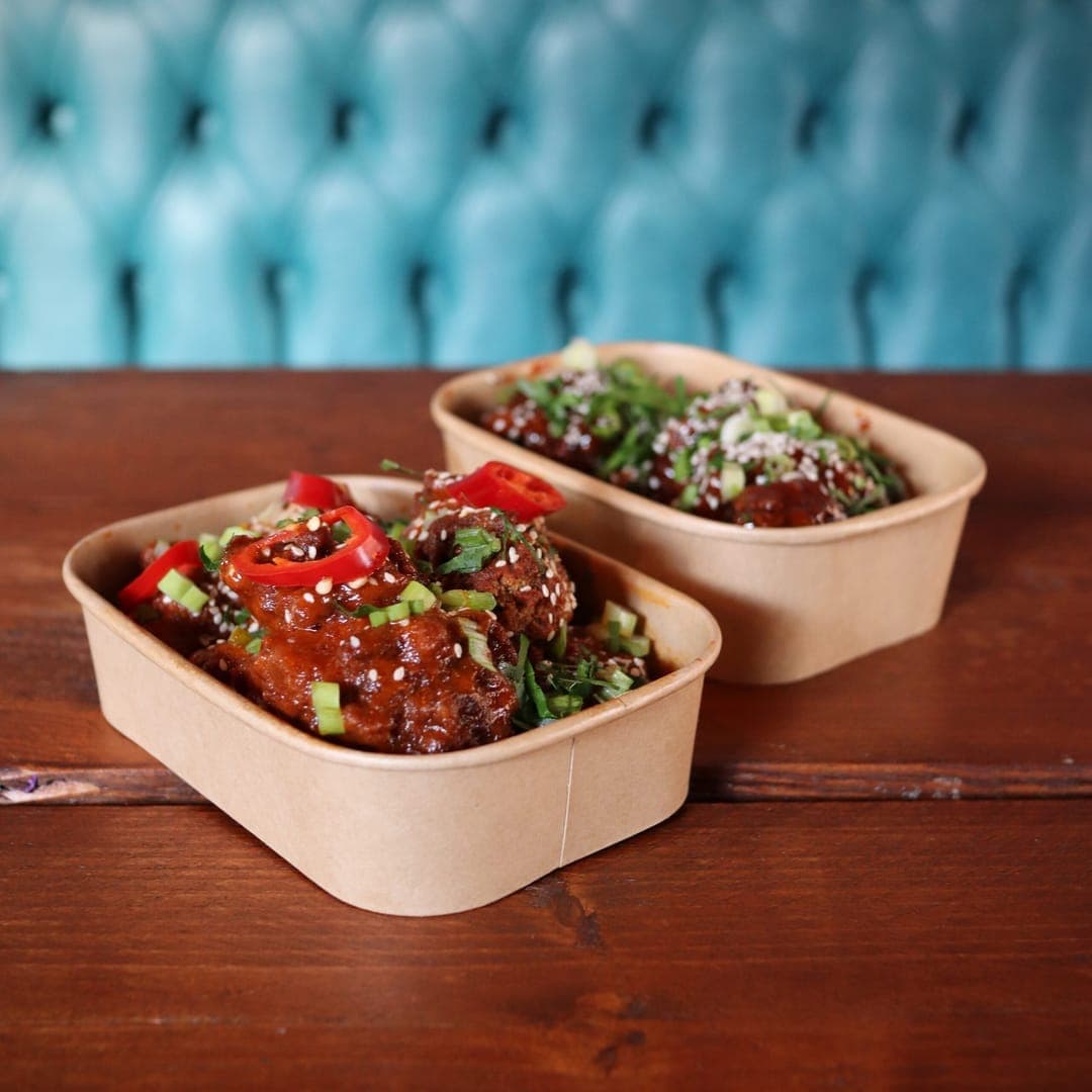 Two paper trays of sticky glazed chicken wings garnished with red chilli, spring onion and sesame seeds on a wooden table