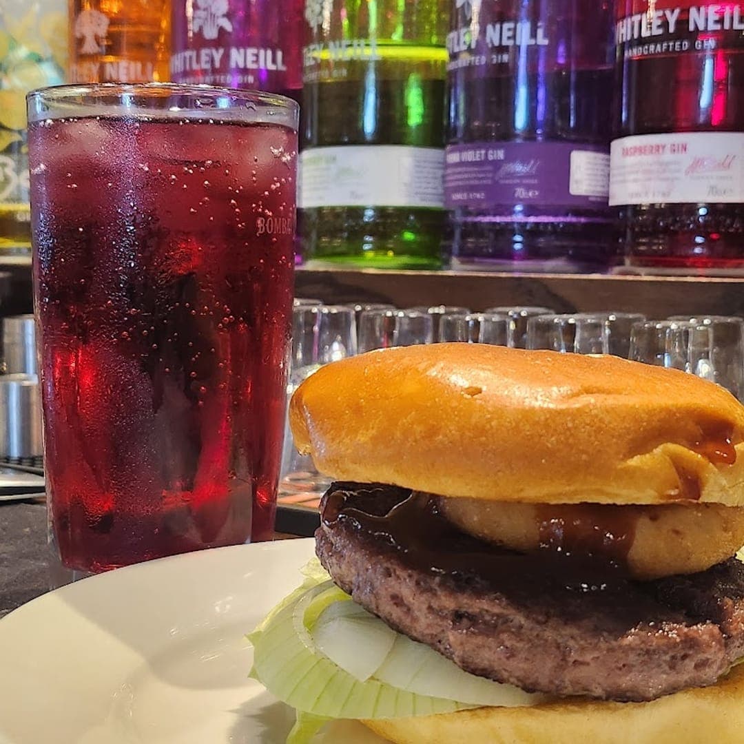 A burger on a white plate with a red cocktail and a colourful gin bottle shelf behind the bar at The Orchard