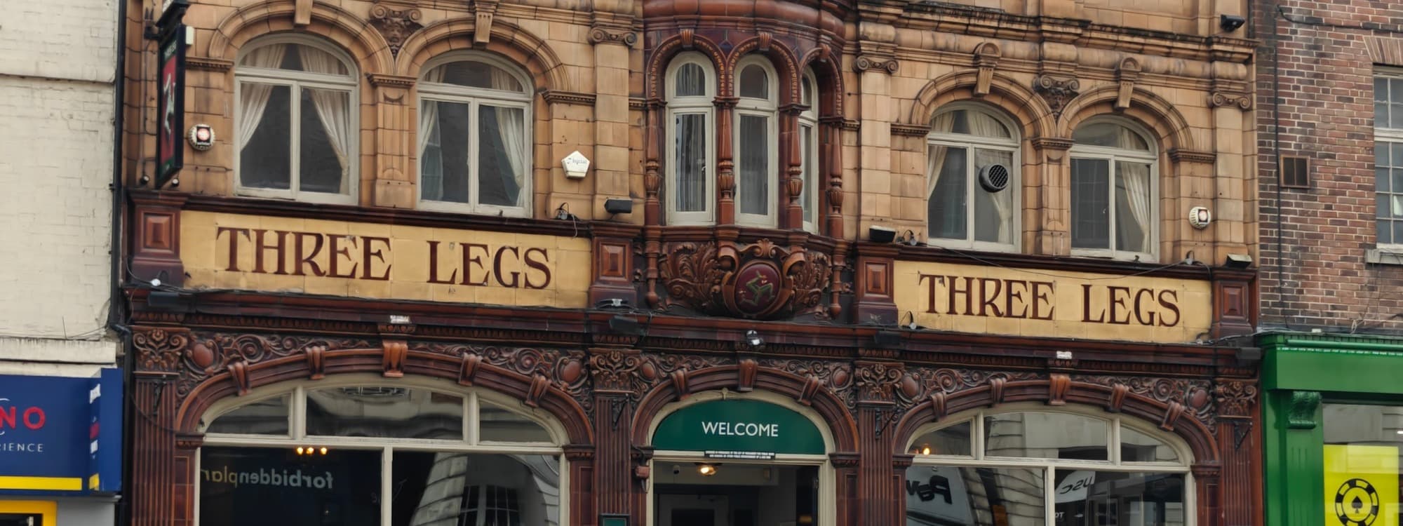 The carved Victorian facade of Three Legs pub on The Headrow, Leeds, with two gold-lettered THREE LEGS bays flanking a central triskelion medallion.