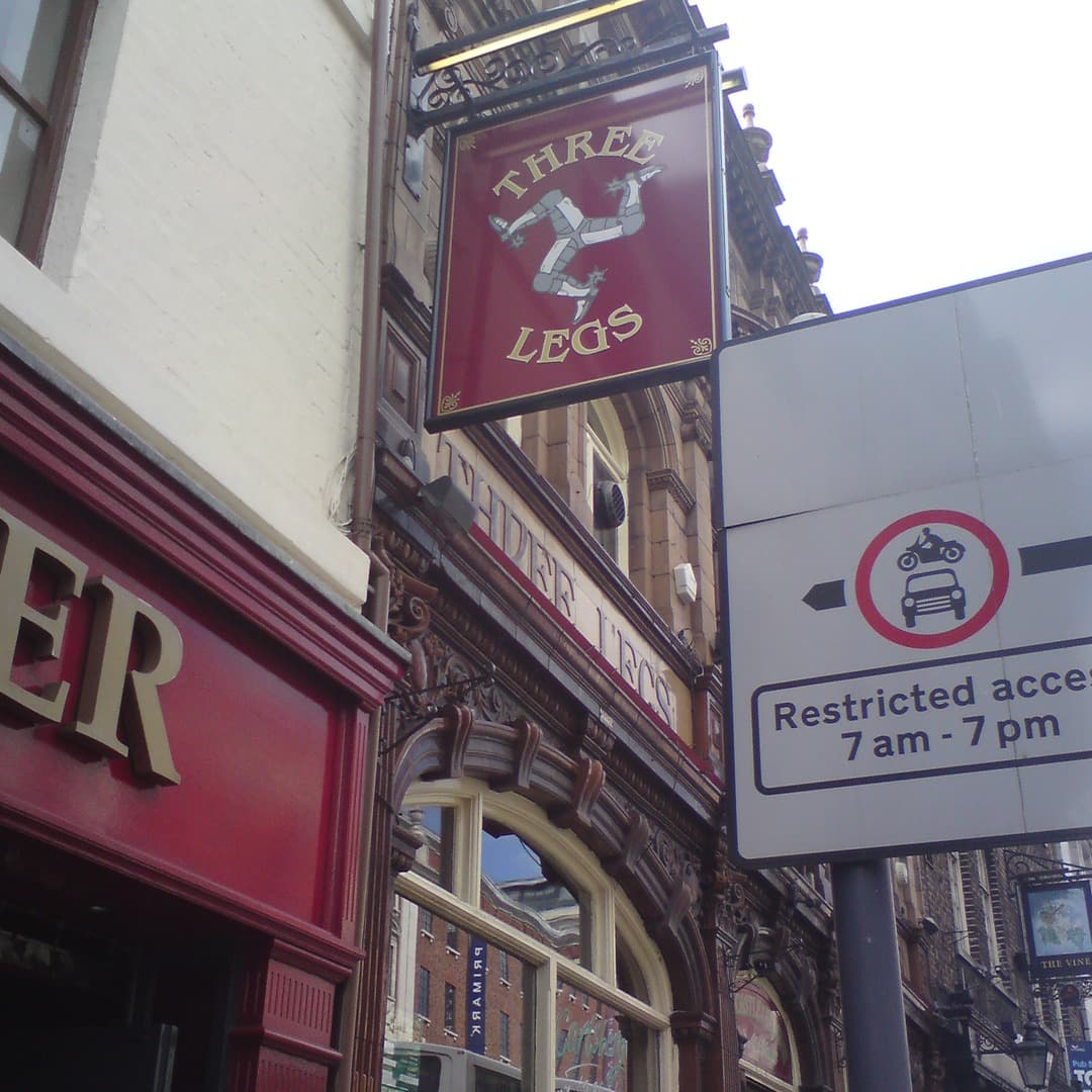 The hanging Three Legs pub sign on its ornate ironwork bracket on The Headrow, Leeds. The maroon sign shows the silver triskelion of the Isle of Man with gold Three Legs lettering.
