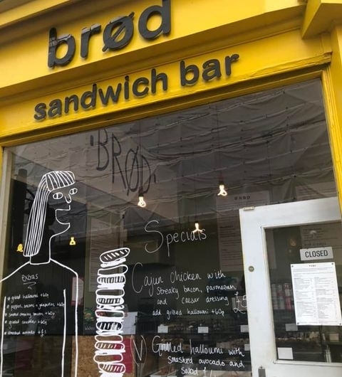Brød Sandwich Bar Yellow Facade Specials Board Exterior view of Brød Sandwich Bar with a bright yellow facade and signage above the entrance. A chalkboard with daily specials is visible in the window, alongside a whimsical drawing of a person. The door has a "Closed" sign.